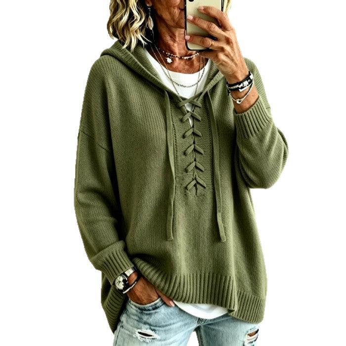 A woman wearing an olive green lace-up hooded sweater over a white top and light blue distressed jeans, holding a phone to take a mirror selfie against a plain white background.