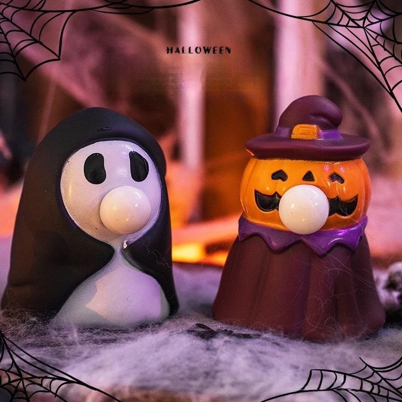 Two small Halloween-themed figurines on a cobwebbed surface: a ghost-like figure in black and white on the left, and a jack-o'-lantern figure in orange, purple, and black on the right. The background is dim and warm-toned with blurred vertical shapes, spiderweb borders, and the word HALLOWEEN at the top.