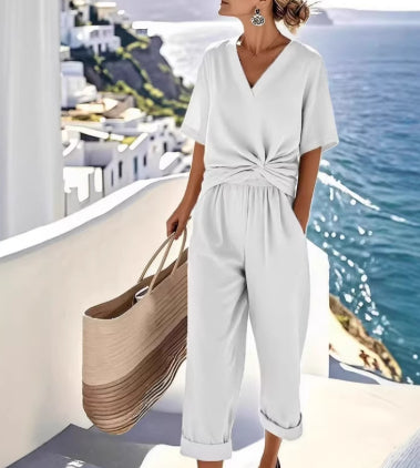 Woman in a white jumpsuit holding a large straw bag on a balcony with ocean view