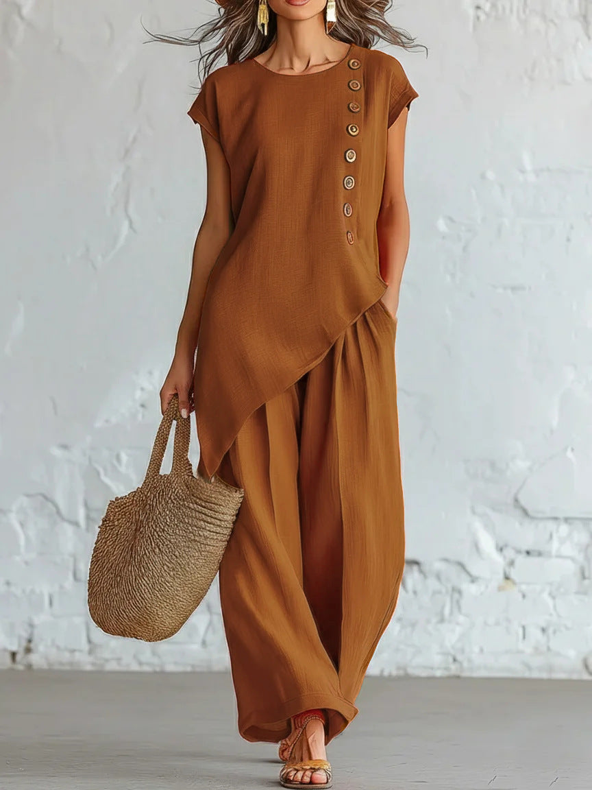 Woman wearing a brown outfit with wide-leg pants and holding a straw bag against a white brick wall.