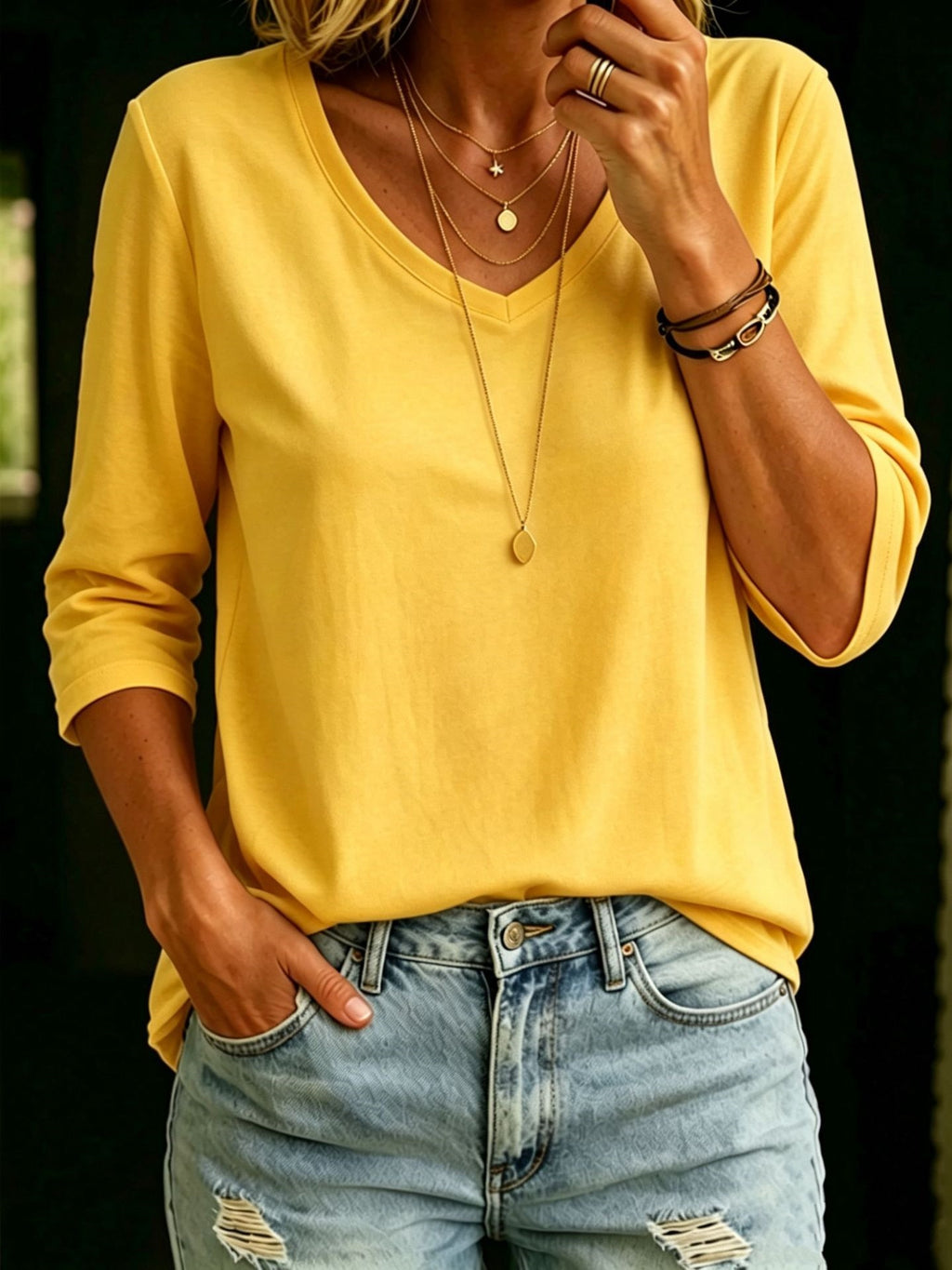 A woman wearing a yellow V-neck long-sleeve top and light blue distressed jeans, accessorized with layered gold necklaces and bracelets.