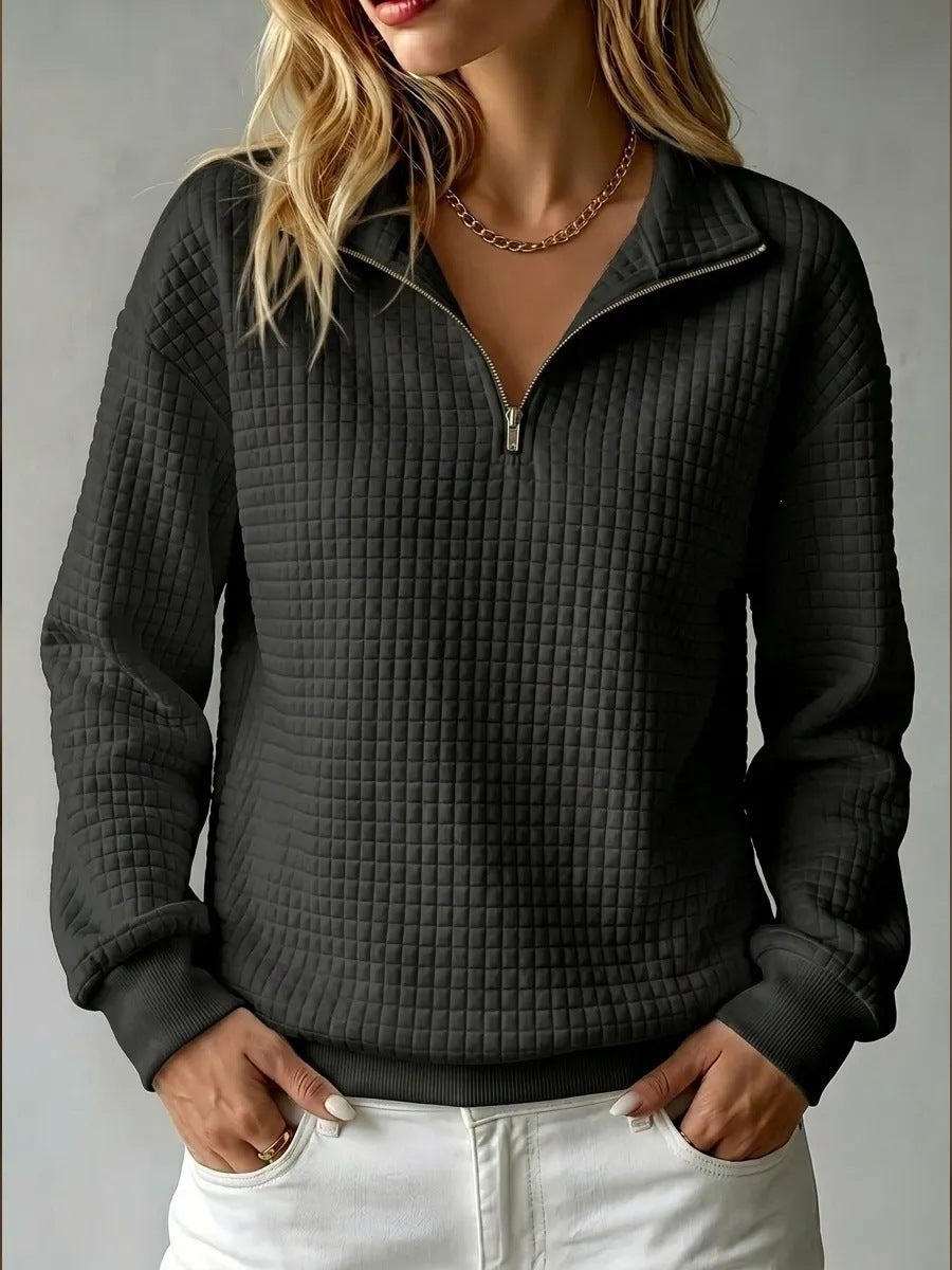A woman wearing a dark gray waffle-textured quarter-zip pullover sweater with a stand collar, paired with white pants, shown against a neutral gray background.