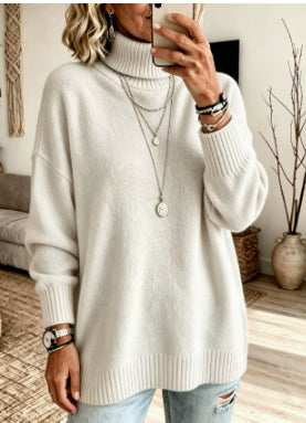 A woman wearing an oversized cream-colored ribbed turtleneck sweater, styled with layered silver necklaces, bracelets, and distressed light-wash jeans, taking a mirror selfie in a cozy living room setting.