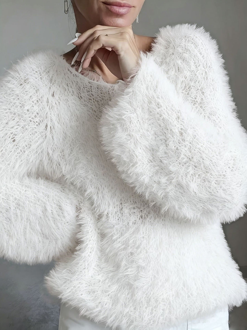 A close-up of a person wearing a fluffy, oversized white knit sweater with voluminous long sleeves and a crew neckline, paired with white pants against a neutral gray background.