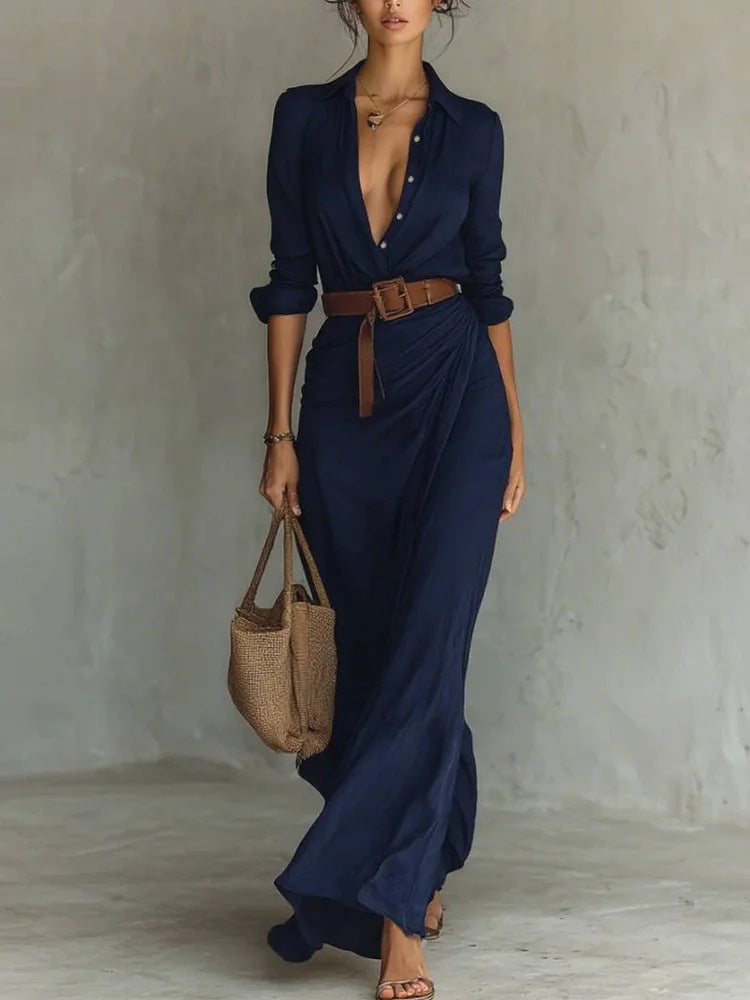 Woman wearing a long navy dress with a brown belt and holding a woven bag against a plain background