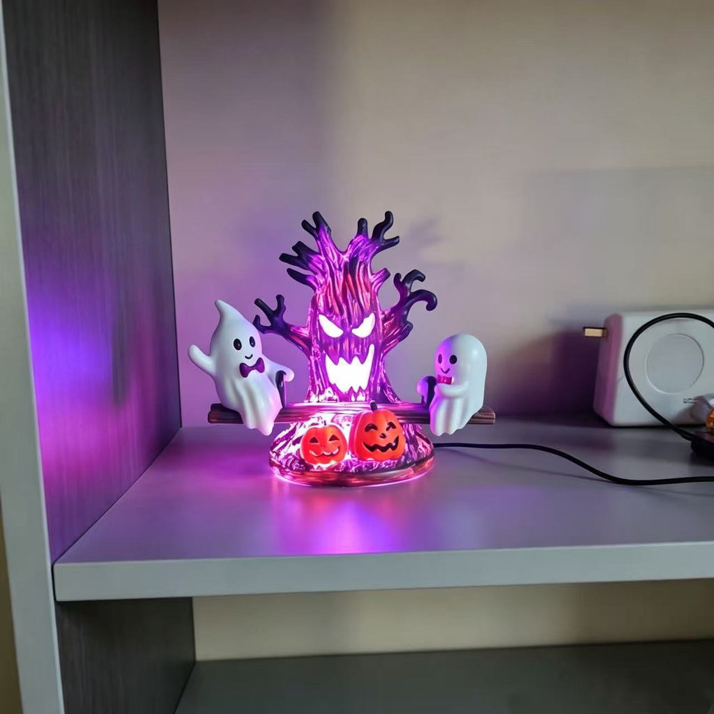A Halloween-themed LED light display featuring a glowing purple tree, two white ghosts, and two jack-o'-lanterns on a clear base, placed on a shelf in a home setting.
