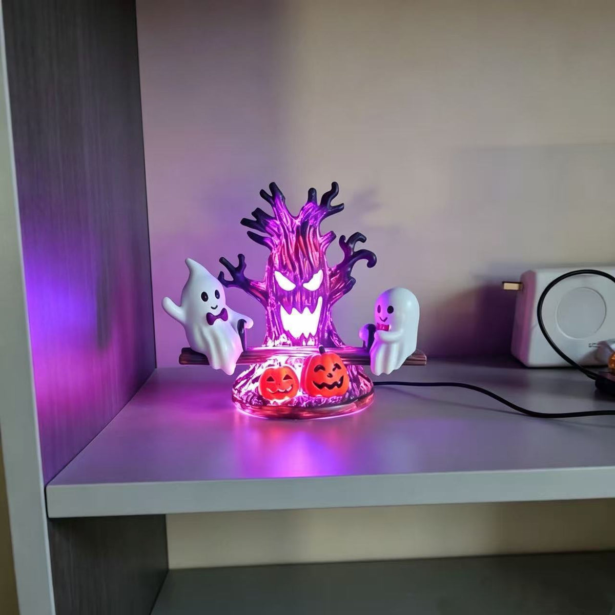 A Halloween-themed LED light display featuring a glowing purple tree, two white ghosts, and two jack-o'-lanterns on a clear base, placed on a shelf in a home setting.