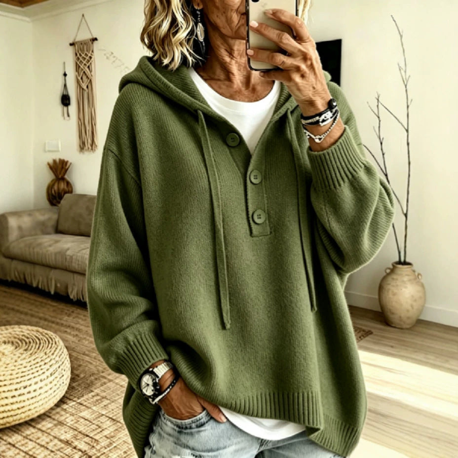 A woman takes a mirror selfie wearing an oversized olive green knit hooded sweater with a buttoned placket over a white top, in a bright living room with a couch, woven rug, and vase.