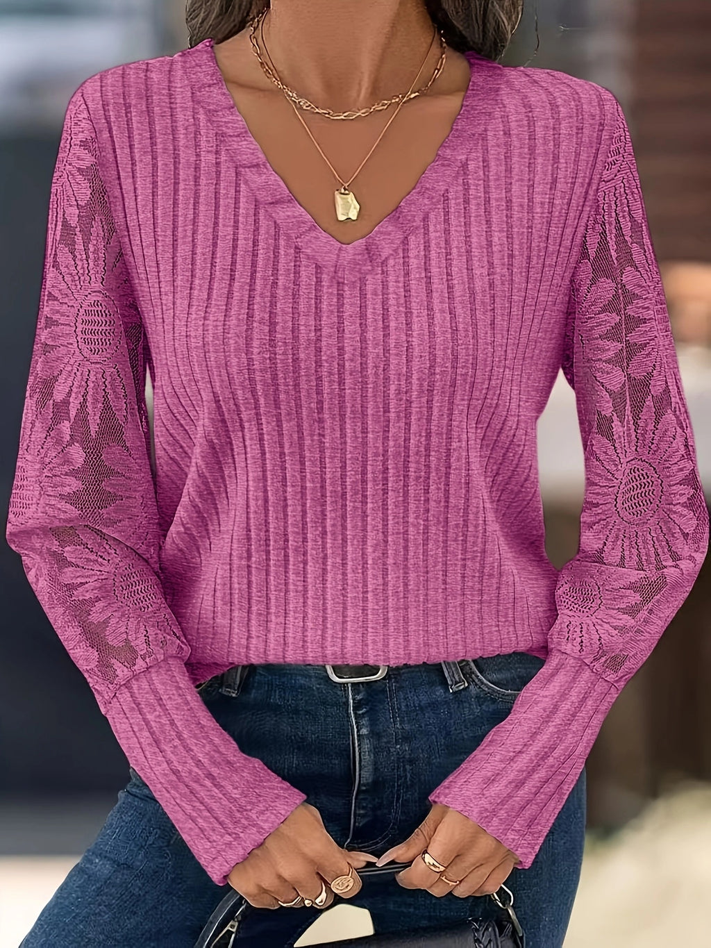 A woman wearing a pink ribbed long-sleeve V-neck sweater with floral lace sleeves, paired with blue jeans and layered gold necklaces.