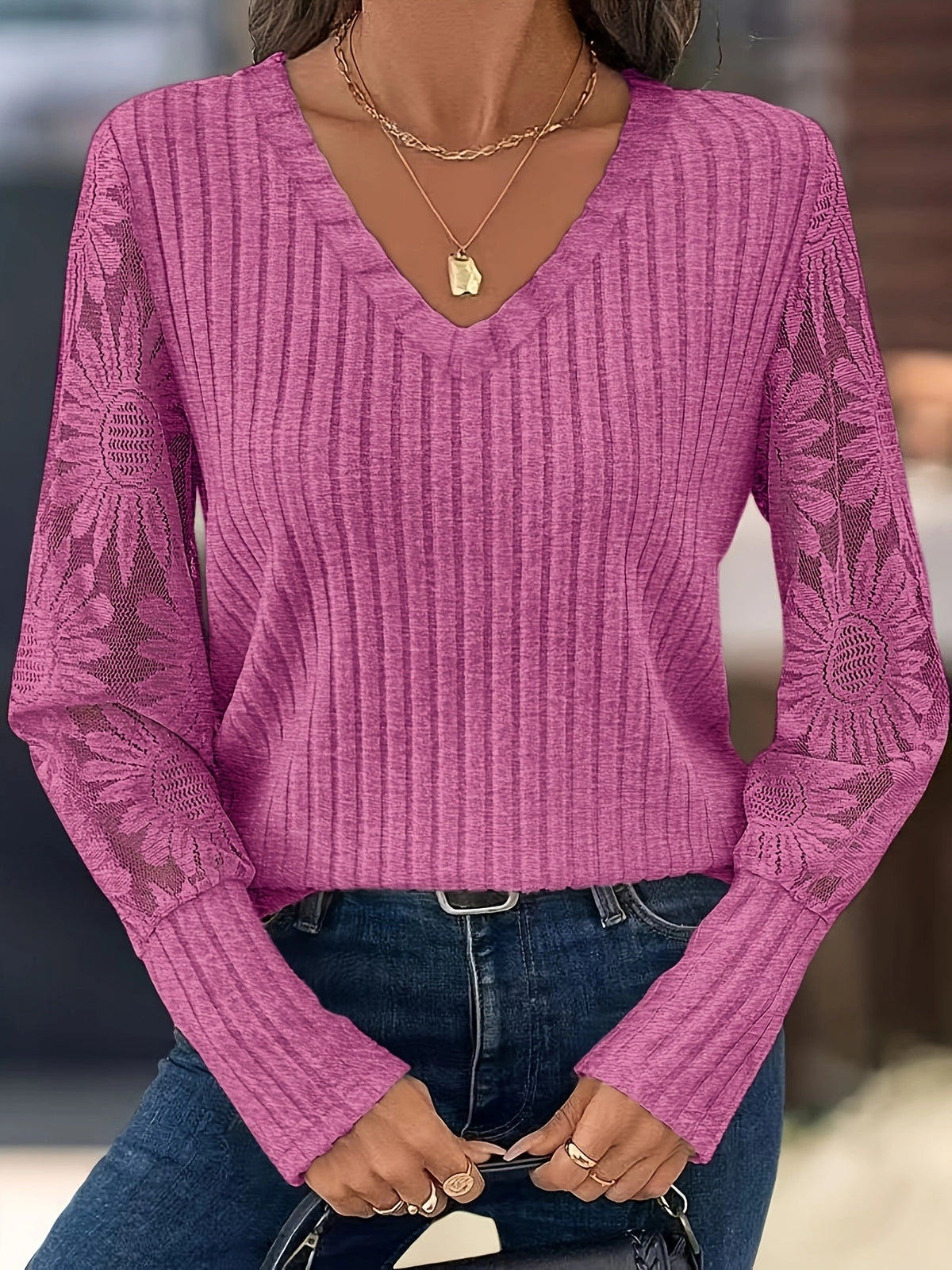 A woman wearing a pink ribbed long-sleeve V-neck sweater with floral lace sleeves, paired with blue jeans and layered gold necklaces.
