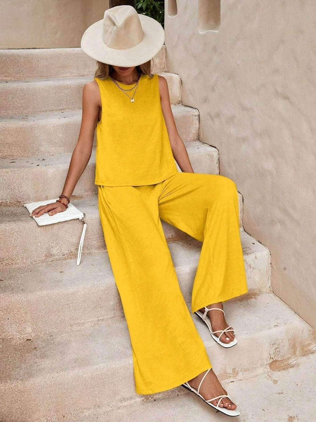 Woman wearing yellow sleeveless top and wide-leg pants, accessorized with a beige hat, sitting on stone stairs, ZA-Zola fashion style.