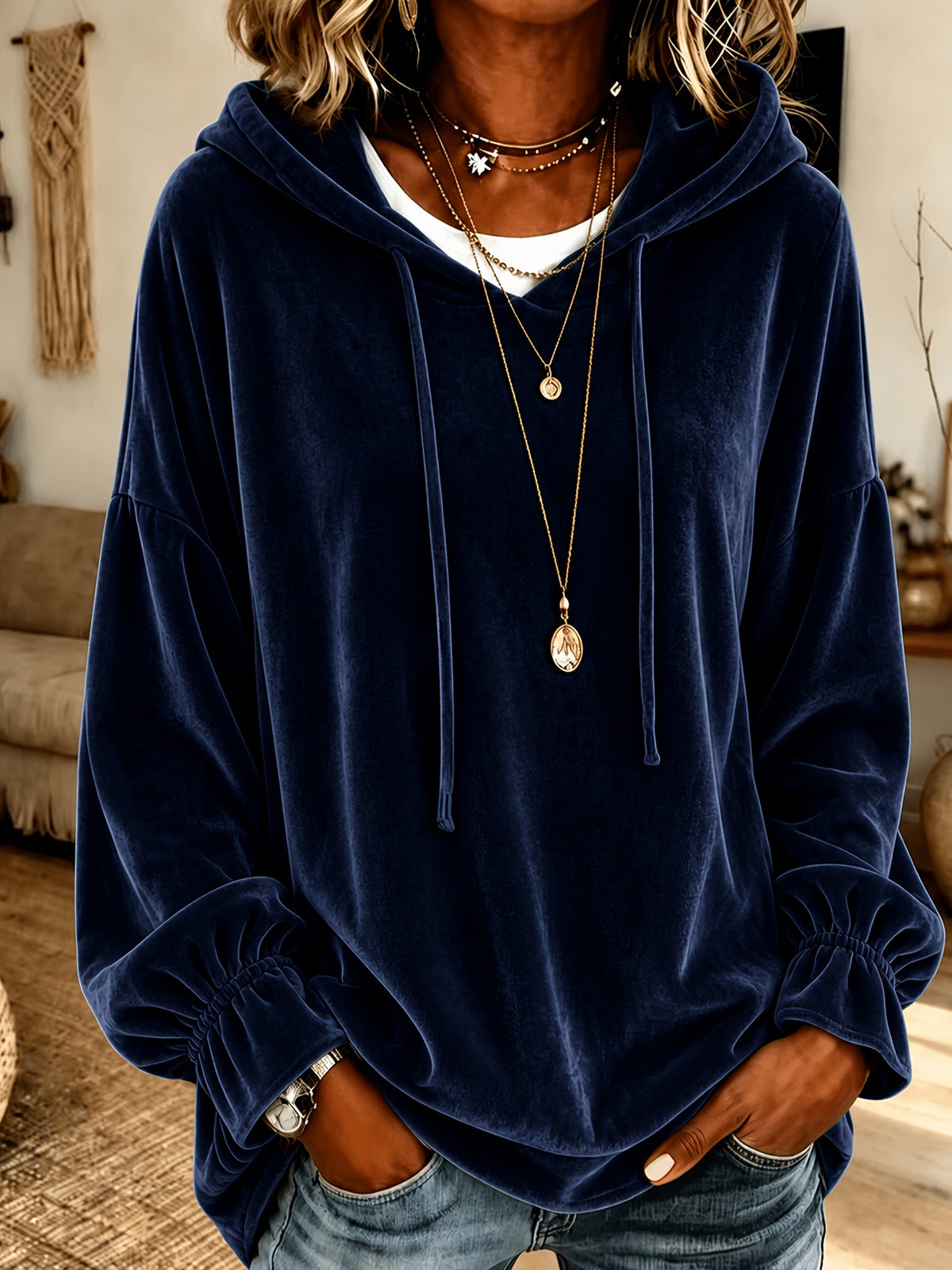 A person wearing a navy blue velvet pullover hoodie with drawstrings and elasticized cuffs, layered over a white undershirt and accessorized with layered necklaces, in a home interior setting.