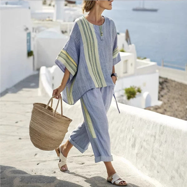 Woman in a light blue outfit with green stripes walking on a sunlit path with white buildings and blue sea in the background.
