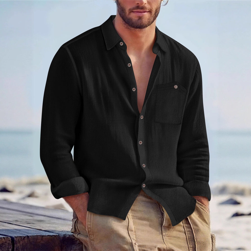 Man wearing a black shirt with rolled-up sleeves and beige pants by the beach.