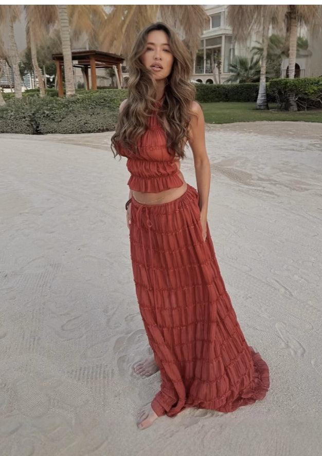 Woman in a rust-colored outfit standing on a sandy surface with greenery in the background