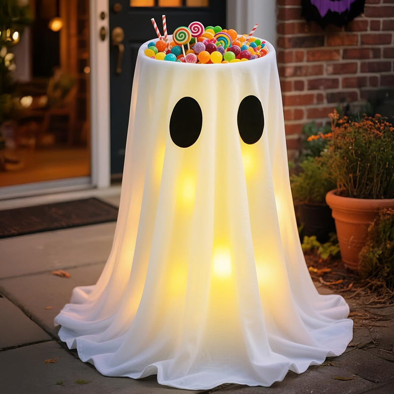 A Halloween ghost-shaped table covered in a white draped cloth with black oval eyes and glowing warm lights inside, topped with colorful lollipops and candy, set outdoors on a front porch.