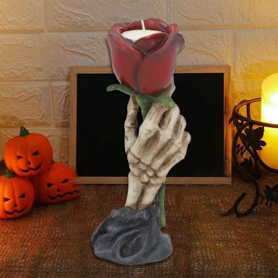 A Halloween-themed candle holder shaped like a skeletal hand holding a red rose with a white candle inside, set on a tabletop with jack-o'-lanterns and a warm light in the background.