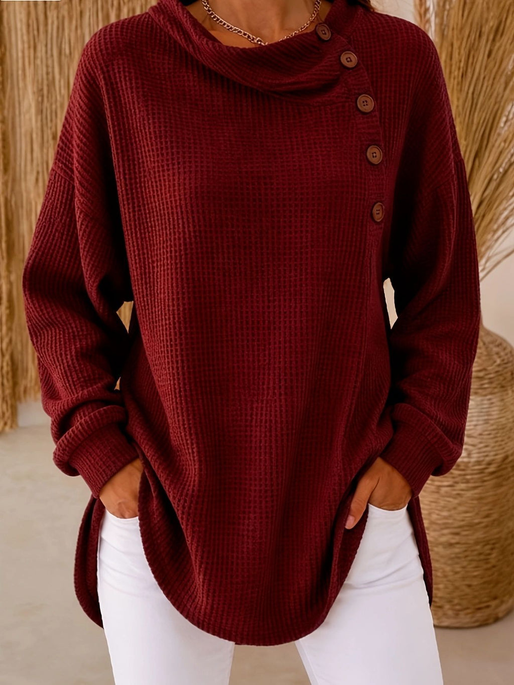 A woman wearing a burgundy waffle-knit long-sleeve sweater with an asymmetrical buttoned cowl neckline and white pants, standing indoors with hands in pockets.