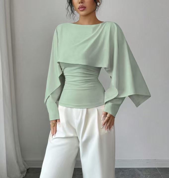 A woman wearing a sage green draped long-sleeve top with white wide-leg pants, standing against a neutral gray wall.