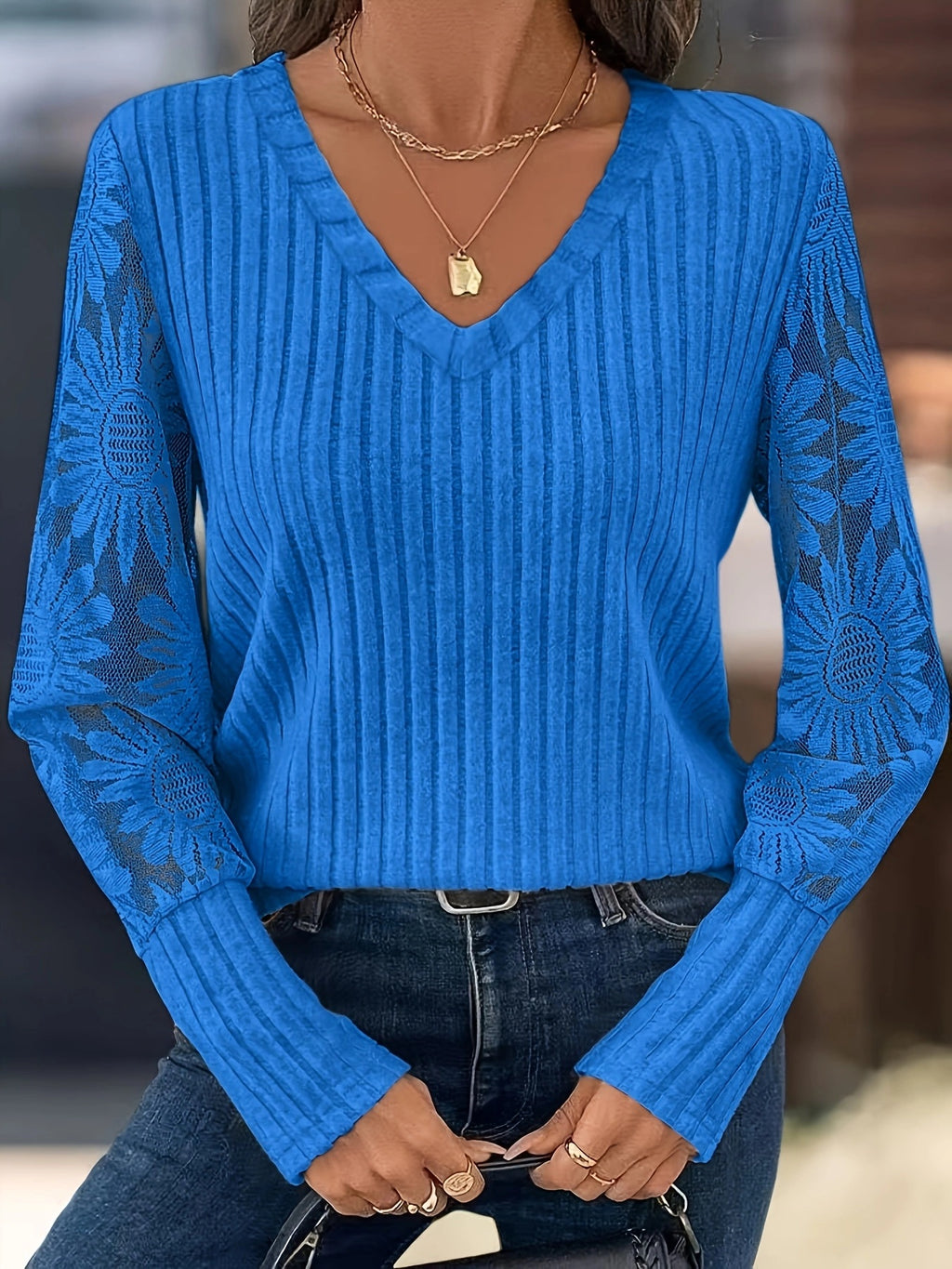 A woman wearing a bright blue ribbed knit sweater with sheer floral lace sleeves, paired with dark blue jeans and layered gold necklaces.