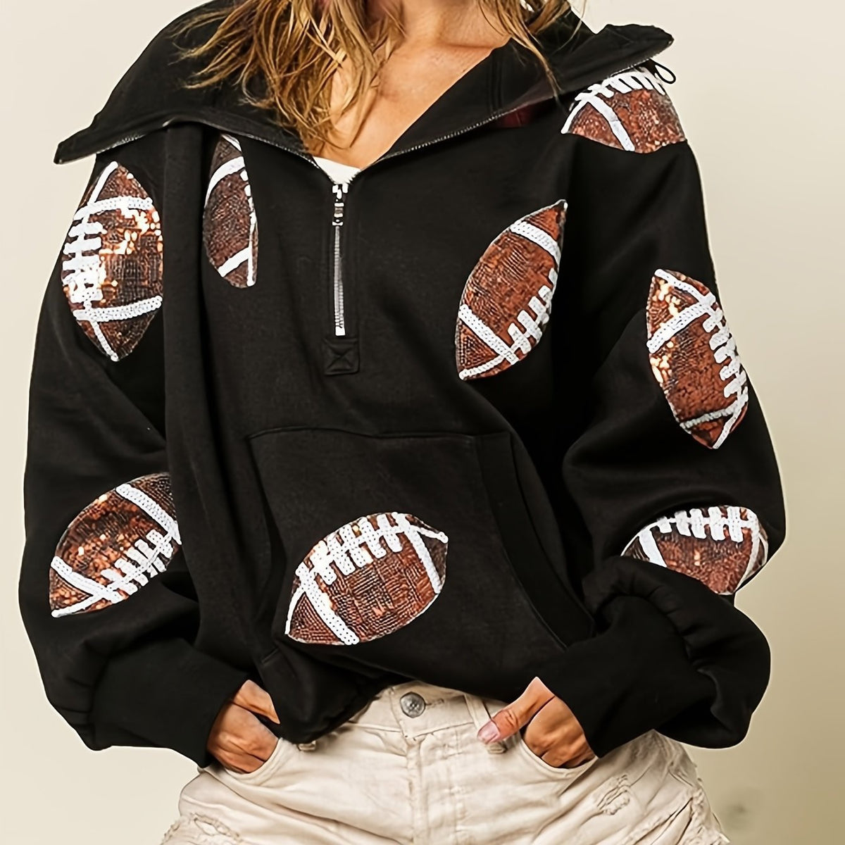 A woman wearing a black pullover hoodie with sequined football patches on the front and sleeves, paired with light beige pants, against a plain beige background.