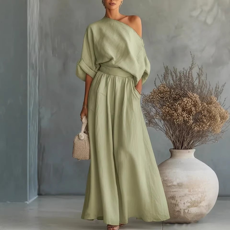 Woman in a green dress holding a small bag, standing next to a large vase with dried plants against a gray wall.