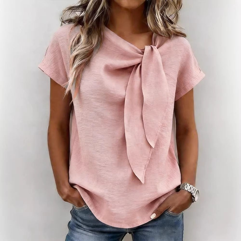 A woman wearing a light pink short-sleeve blouse with a large front tie detail, paired with blue jeans, shown against a plain light gray background.