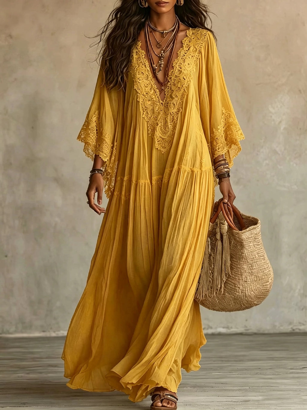 A woman wearing a flowing mustard yellow lace-trim maxi dress with wide sleeves, holding a woven straw tote bag against a textured neutral wall.