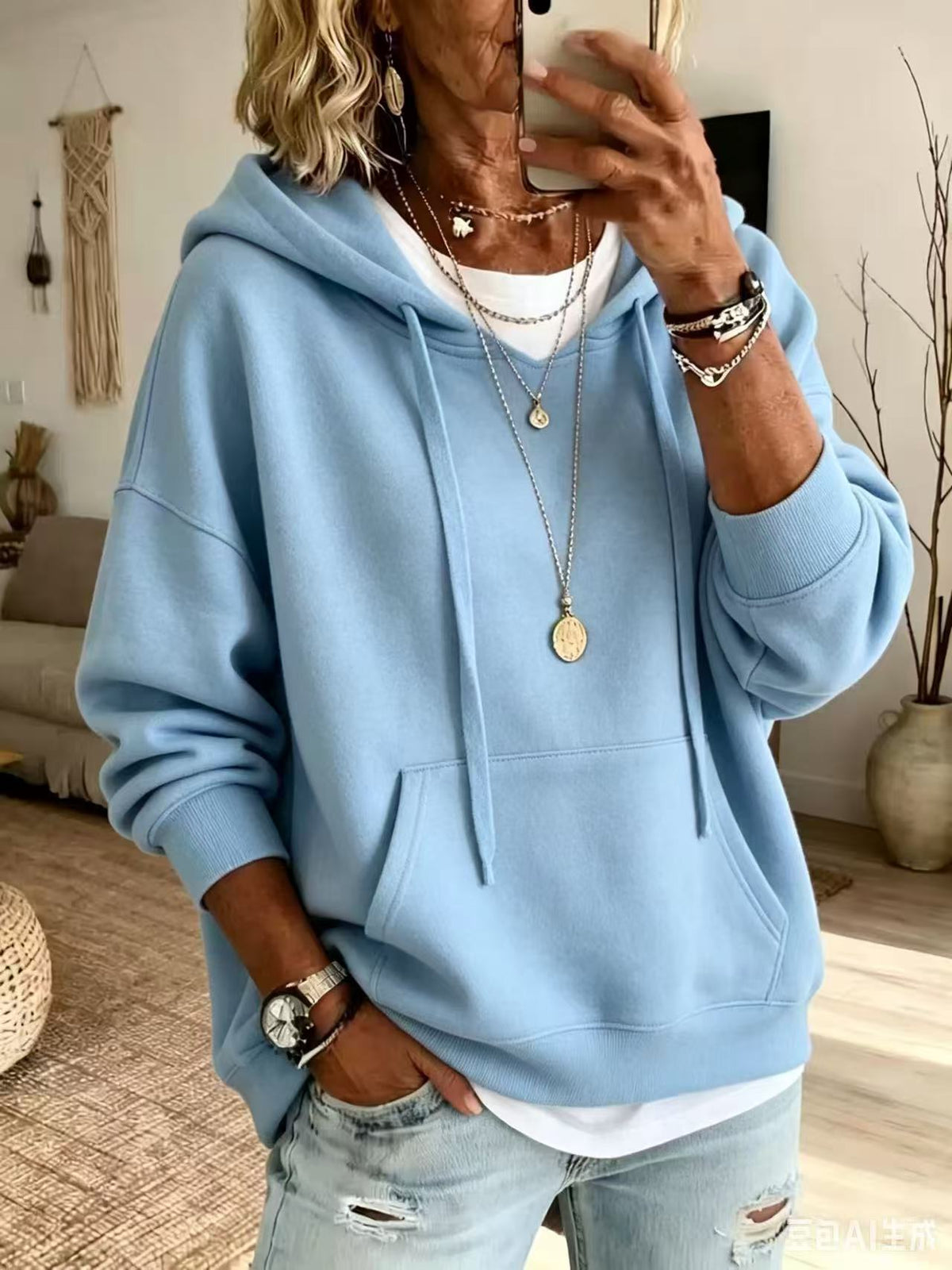 A person taking a mirror selfie wearing a light blue oversized pullover hoodie with drawstrings, a white undershirt, and light-wash distressed jeans. The setting is a cozy indoor room with neutral decor.