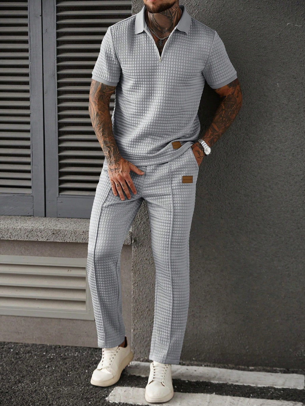 Man wearing a checkered outfit with a gray shirt and pants, standing against a gray wall.