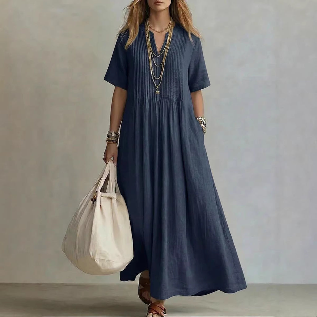 A woman wearing a navy blue linen maxi dress with short sleeves, a smocked chest, and side pockets, holding a large cream canvas tote bag. She is accessorized with layered gold necklaces and bracelets, standing against a neutral textured gray wall.