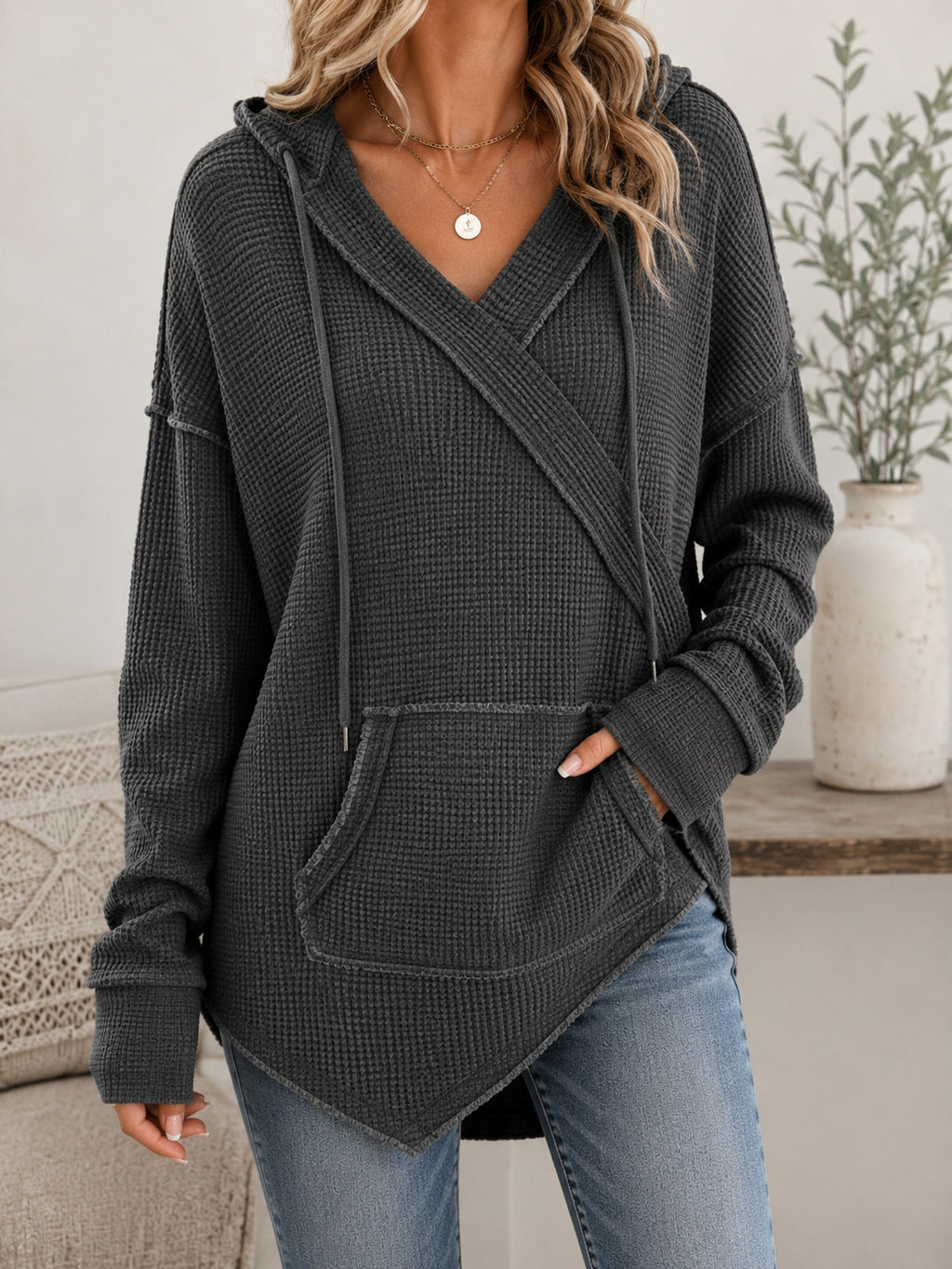 A woman wearing a dark gray waffle-knit hooded sweater with a wrap-style front and kangaroo pockets, paired with blue jeans in a softly lit indoor setting.