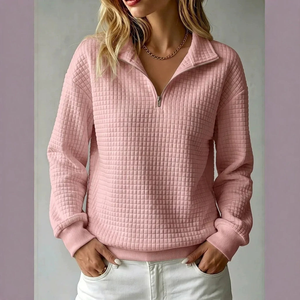 A woman wearing a pink waffle-textured quarter-zip pullover sweater with a stand collar, paired with white pants, shown against a neutral studio background.