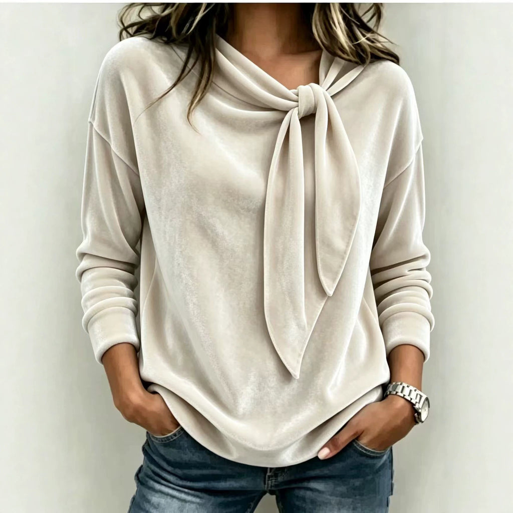 A woman wearing a light beige long-sleeve knit top with a large tie-neck detail, paired with blue jeans, standing against a plain light gray wall.