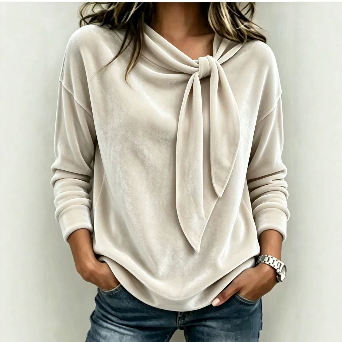A woman wearing a light beige long-sleeve knit top with a large tie-neck detail, paired with blue jeans, standing against a plain light gray wall.