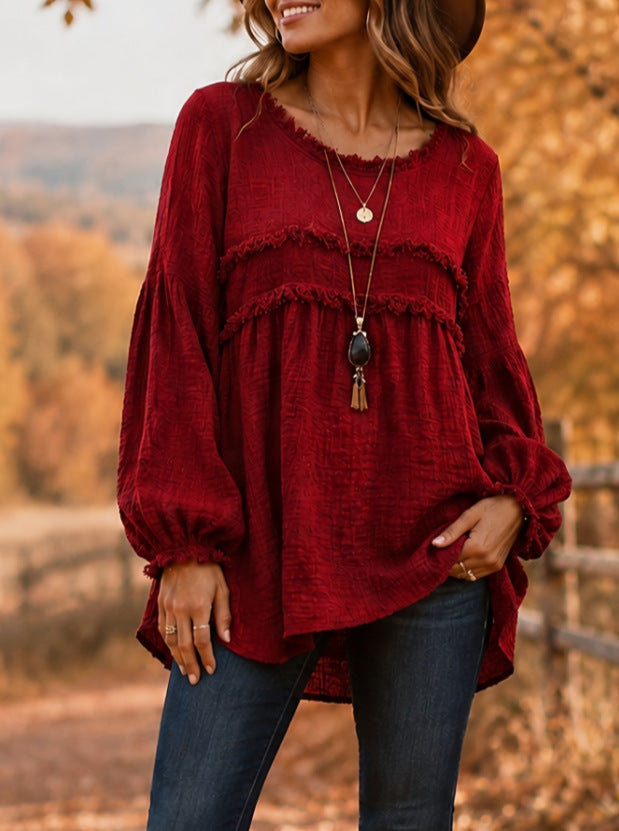 A woman wearing a long-sleeve, deep red textured blouse with a ruffled yoke and gathered cuffs, paired with dark blue jeans, set against an outdoor autumn background.