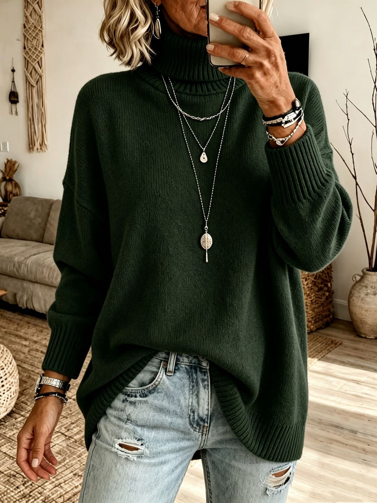 A woman wearing a dark forest green ribbed turtleneck sweater and light-wash distressed jeans, taking a mirror selfie in a cozy living room setting.
