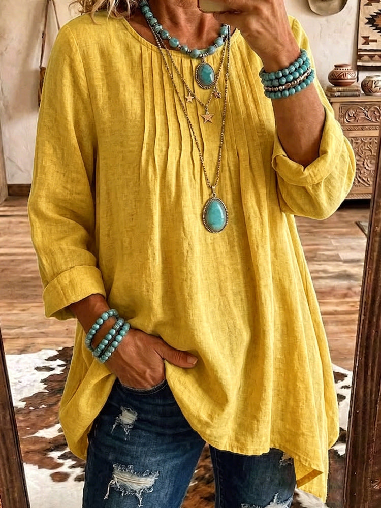 A woman wearing a loose, yellow linen-blend tunic top with long sleeves, paired with distressed blue jeans and layered turquoise jewelry, taking a mirror selfie in a rustic room.