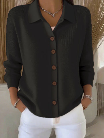 Person wearing a black button-up shirt with brown buttons and white pants indoors.