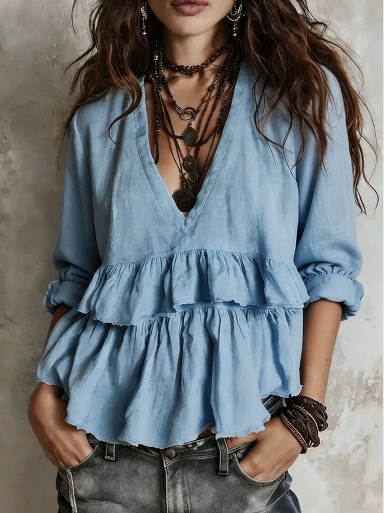 A woman wearing a light blue tiered ruffle blouse with a deep V-neck, layered with multiple dark necklaces and bracelets, paired with gray jeans against a textured gray wall.