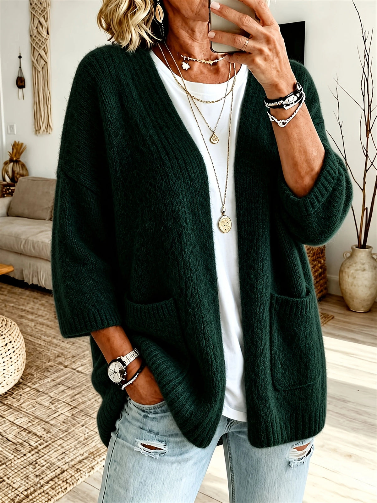 A woman wearing a dark forest green chunky knit cardigan over a white t-shirt and light-wash distressed jeans, shown in a cozy indoor room setting.