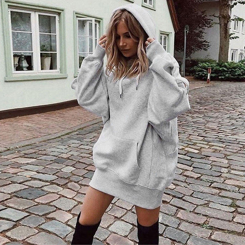 A woman wearing an oversized light gray heather pullover hoodie with a drawstring hood and front kangaroo pocket, styled as a mini sweatshirt, standing on a cobblestone street in front of a light-colored building.