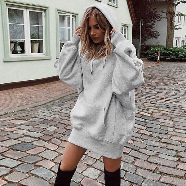 A woman wearing an oversized light gray heather pullover hoodie with a drawstring hood and front kangaroo pocket, styled as a mini sweatshirt, standing on a cobblestone street in front of a light-colored building.