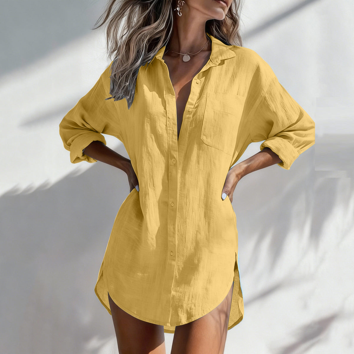 A woman wearing a loose, oversized yellow button-up shirt dress with rolled sleeves and side slits, standing against a white wall with soft shadow patterns.