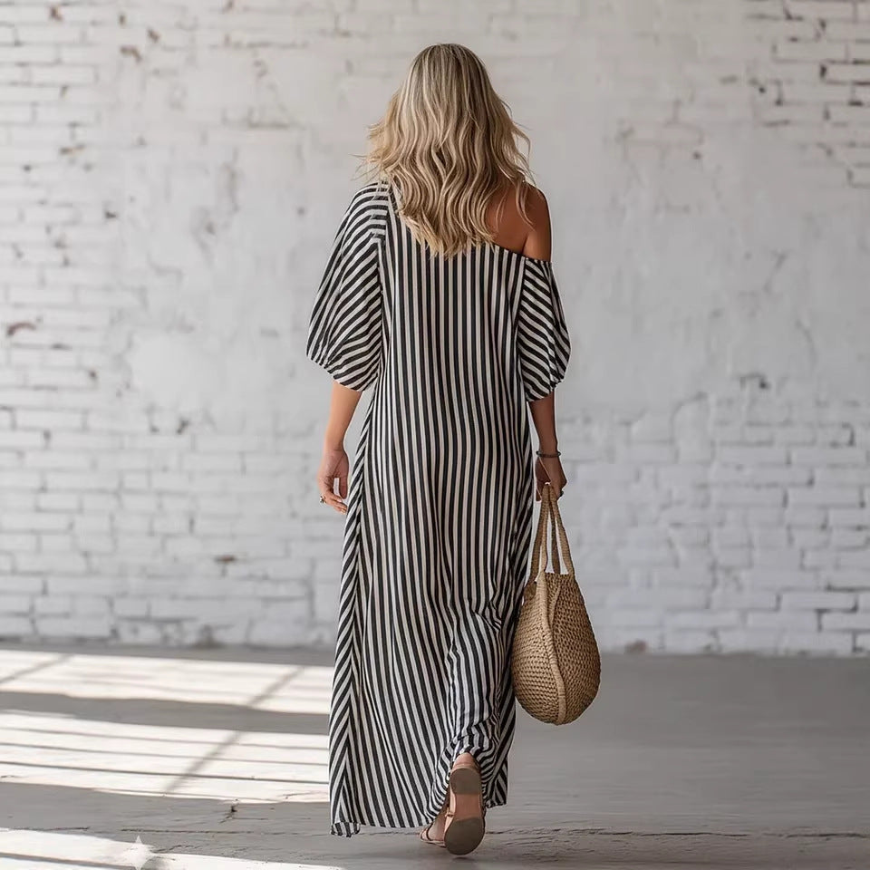 Woman in a black and white striped dress holding a woven bag against a white brick wall.