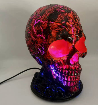 A decorative skull-shaped lamp with a black base, featuring glowing RGB LED lighting that shifts between red, pink, purple, and blue. The skull has a textured, cracked metallic finish, and a power cord is visible on the left against a neutral gray background.