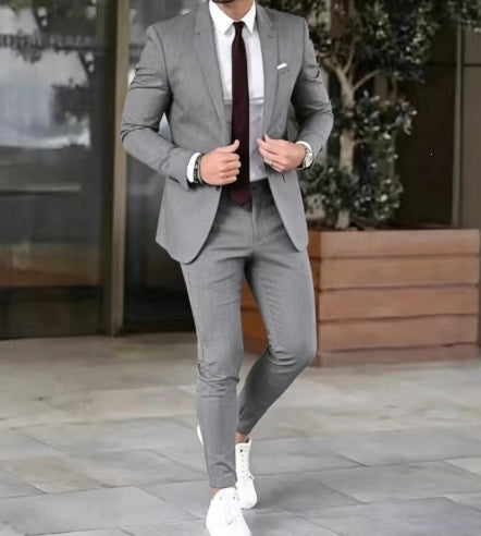 A man wearing a slim-fit gray three-piece suit with a white dress shirt, a dark burgundy tie, and white sneakers, standing in front of a modern building entrance.