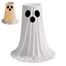 A white ghost-shaped tablecloth with black eyes and mouth details, shown on a table with a small inset of a decorated cake wearing a matching ghost hat.