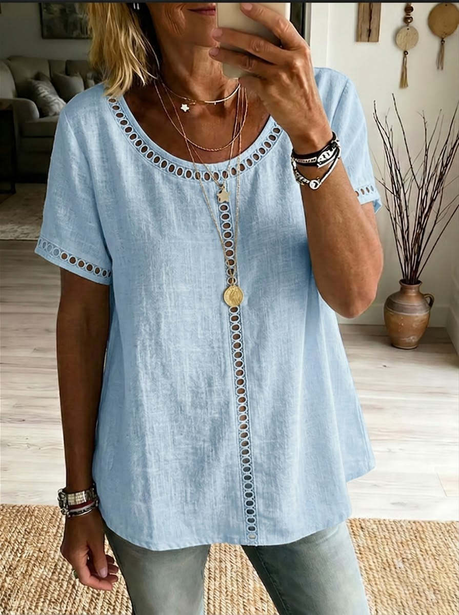 Woman wearing a light blue top with decorative buttons and trim, standing in a room with a couch and decorative items.