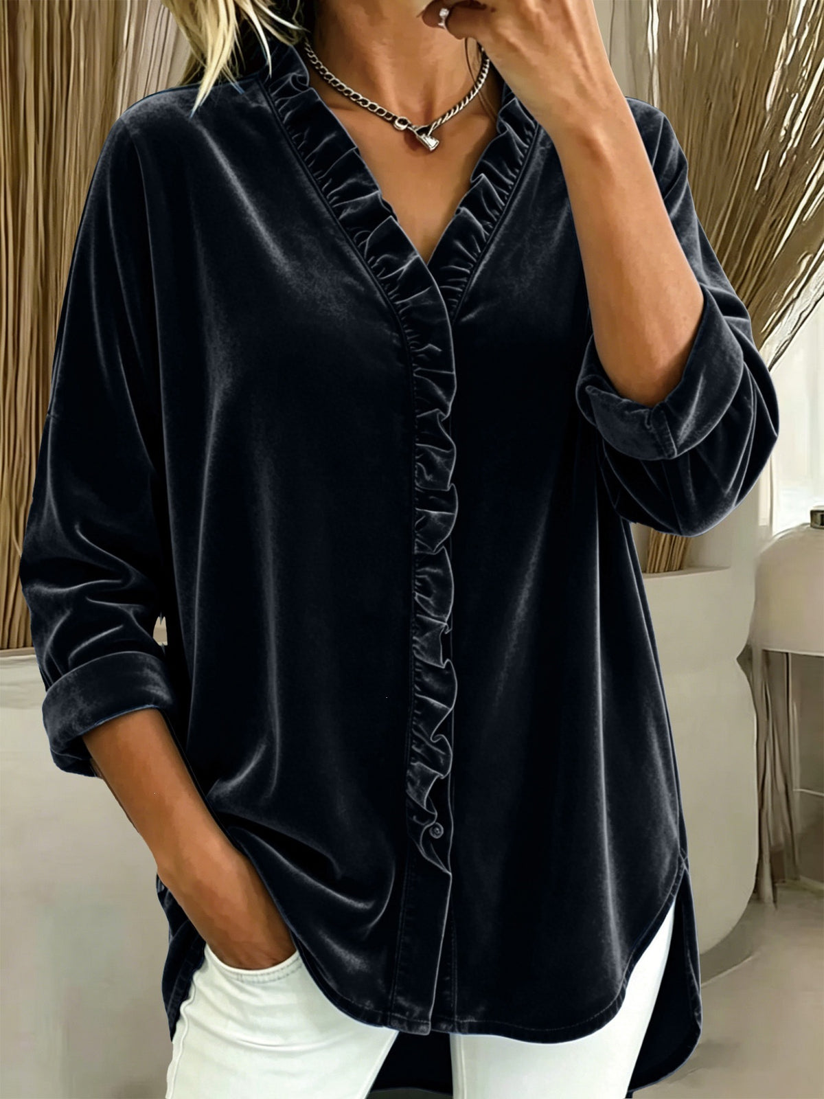 A woman wearing a black velvet long-sleeve blouse with ruffle trim along the V-neck and front opening, paired with white pants in an indoor setting.